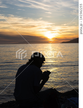 Male adult traveler enjoying a scenic sunset view from the shore, documenting the picturesque seascape during golden hour with a camera Male adult traveler enjoying a scenic sunset view from the shore, documenting the picturesque seascape during golden hour with a camera 141562255