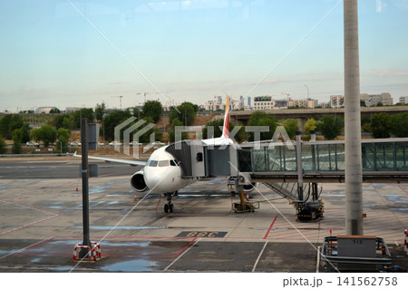 Parked aircraft at jet bridge on airport tarmac seen through terminal window for travel background and gate boarding scene with runway view Parked aircraft at jet bridge on airport tarmac seen through terminal window for travel background and gate boarding scene with runway view 141562758