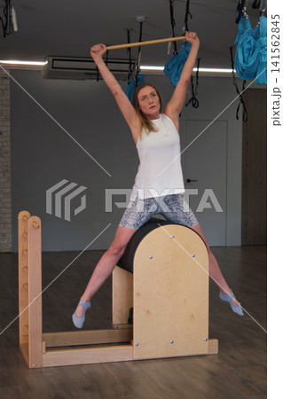 A female client balances on a ladder barrel in a wide seated stretch while reaching upward with a wooden stick. The photo suits fitness industry themes of service, mobility, safety, and wellbeing. 141562845