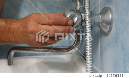 A man's hand adjusts or repairs a chrome faucet valve in a bathroom. The long spout and the edge of a white sink are visible below, and a shower hose is visible to the right. A man's hand adjusts or repairs a chrome faucet valve in a bathroom. The long spout and the edge of a white sink are visible below, and a shower hose is visible to the right. 141563314