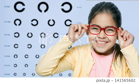 Happy little girl wearing bright red glasses frame during a pediatric vision diagnostic consultation. Happy little girl wearing bright red glasses frame during a pediatric vision diagnostic consultation. 141565682