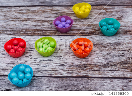 Colorful balls sorted into silicone cups lie on rustic wooden background. Bright plastic spheres promote early child development through color matching activity. Educational toy set fosters skills. Colorful balls sorted into silicone cups lie on rustic wooden background. Bright plastic spheres promote early child development through color matching activity. Educational toy set fosters skills. 141566300