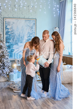 a family in elegant light-blue and white attire opening a gift in a magical winter wonderland a family in elegant light-blue and white attire opening a gift in a magical winter wonderland 141566640