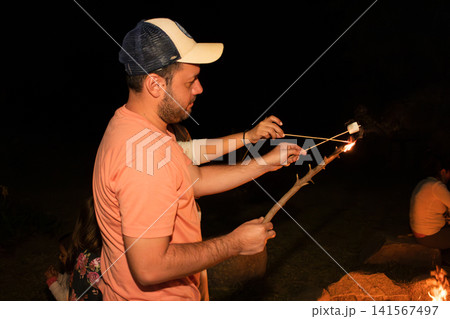 Man roasting marshmallows over night campfire with family Man roasting marshmallows over night campfire with family 141567497
