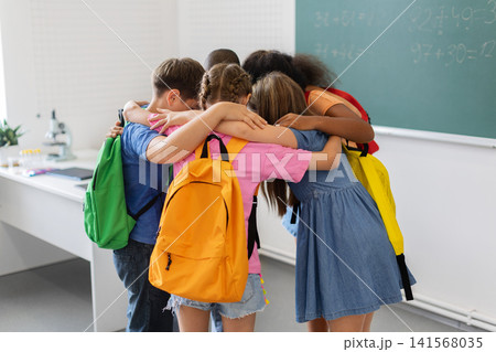 Diverse schoolchildren wearing colorful backpacks sharing close group hug in the classroom 141568035