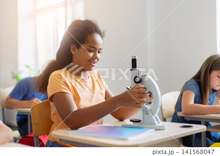 Smiling black schoolgirl adjusting microscope during engaging science lesson at her desk 141568047