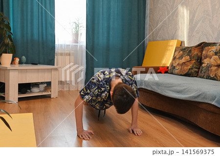 Boy does an exercise on a yellow mat in a room with a green plant nearby during the day 141569375