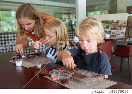 Kids and their mother participate in a chocolate making workshop in Bali, decorating sweets with toppings. A fun and educational family activity during tropical holidays. 141570031