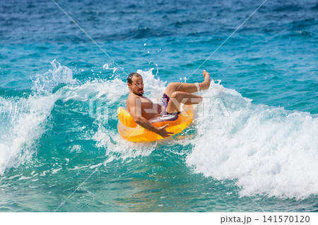 Man falling off inflatable ring while playing in ocean waves. Fun, energetic beach moment captured mid-splash. Ideal for summer vacation, tropical holiday, joy, active water recreation, sea adventure 141570120
