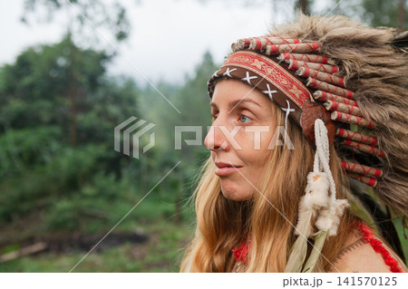 Portrait of a young woman wearing traditional Indian feathered costume. Image was taken outdoors in a forest like setting, evoking a cultural or educational experience for children. Portrait of a young woman wearing traditional Indian feathered costume. Image was taken outdoors in a forest like setting, evoking a cultural or educational experience for children. 141570125