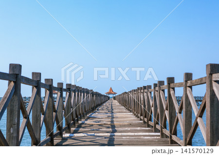 Long wooden pier with traditional gazebos - bale stretches over calm turquoise sea under clear blue sky. Perfect place for viewing amazing landscape. Scenery for travel, resort, summer vacation Long wooden pier with traditional gazebos - bale stretches over calm turquoise sea under clear blue sky. Perfect place for viewing amazing landscape. Scenery for travel, resort, summer vacation 141570129