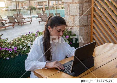 Young Female Freelancer Typing on Tablet Keyboard at Outdoor Resort Cafe 141570341
