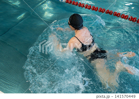 Young woman in black swimsuit swimming in the pool, feet in water, resting near lane divider inside modern swimming facility with wet hair, calm posture and natural light Young woman in black swimsuit swimming in the pool, feet in water, resting near lane divider inside modern swimming facility with wet hair, calm posture and natural light 141570449