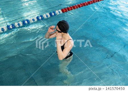 Mature woman adjusting swim goggles in indoor lap pool, wearing swim cap and focused before training, waist-deep in calm water with bright windows and modern wooden ceiling 141570450