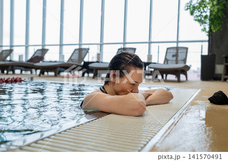 Young person resting beside indoor pool edge with folded arms, wet short hair, calm pensive expression, blue water reflections, tiled deck, and quiet wellness setting 141570491
