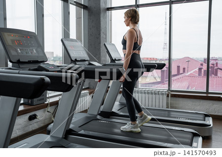 Young woman walking on treadmill in bright modern gym studio with floor-to-ceiling windows and city rooftops outside, surrounded by rows of cardio machines and soft daylight 141570493