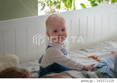 Smiling baby sits beside older brother on bed in cozy children bedroom during warm family moment. Childhood, parenting, sibling bond, home lifestyle, emotional connection, comfort, togetherness, care 141571120