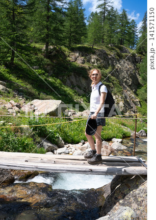 Woman backpacking in an alpine forest setting, crossing a rustic wooden bridge over a flowing mountain stream, enjoying nature and outdoor adventure during summer Woman backpacking in an alpine forest setting, crossing a rustic wooden bridge over a flowing mountain stream, enjoying nature and outdoor adventure during summer 141571150