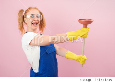 Energetic woman wearing safety glasses, work overalls, and yellow rubber gloves, actively struggling with a drain plunger, showcasing a determined yet surprised expression on a pink background Energetic woman wearing safety glasses, work overalls, and yellow rubber gloves, actively struggling with a drain plunger, showcasing a determined yet surprised expression on a pink background 141571239