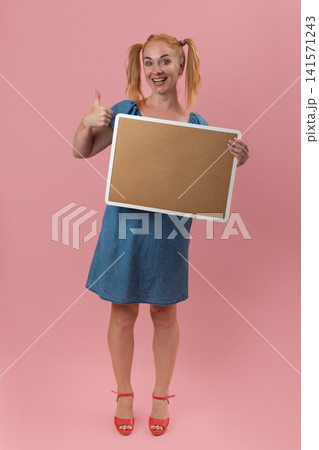 Smiling young woman with red hair in pigtails holding a blank cork board sign offering copy space, giving a thumbs up gesture on a pink background, expressing approval and positivity Smiling young woman with red hair in pigtails holding a blank cork board sign offering copy space, giving a thumbs up gesture on a pink background, expressing approval and positivity 141571243