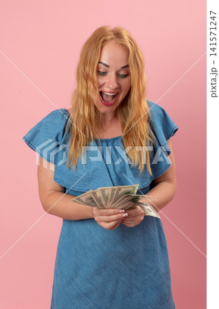Cheerful young woman in a denim dress holding a fan of dollar banknotes, feeling excited and shocked with a wide open mouth expression on a pink background Cheerful young woman in a denim dress holding a fan of dollar banknotes, feeling excited and shocked with a wide open mouth expression on a pink background 141571247