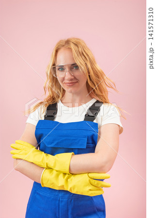 Young woman in protective workwear. Including blue overalls. Yellow gloves. And safety glasses. Standing confidently with crossed arms against a pink background Young woman in protective workwear. Including blue overalls. Yellow gloves. And safety glasses. Standing confidently with crossed arms against a pink background 141571310