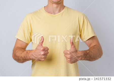 Man in a yellow t shirt presenting two thumbs up, expressing acceptance and agreement, symbolizing approval, success, and positive feedback on a grey background Man in a yellow t shirt presenting two thumbs up, expressing acceptance and agreement, symbolizing approval, success, and positive feedback on a grey background 141571332