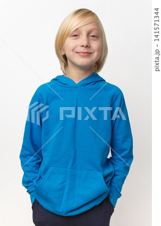 Young blonde boy with hands in his pockets, wearing a bright blue hooded sweatshirt and plain dark pants, smiling confidently while standing against a clean white background Young blonde boy with hands in his pockets, wearing a bright blue hooded sweatshirt and plain dark pants, smiling confidently while standing against a clean white background 141571344