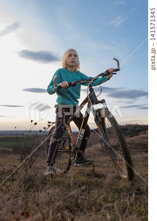 Young boy with blonde hair sitting on a mountain bike on a grassy hill, looking up at the sky, representing concepts of childhood adventure, freedom, and future aspirations Young boy with blonde hair sitting on a mountain bike on a grassy hill, looking up at the sky, representing concepts of childhood adventure, freedom, and future aspirations 141571345
