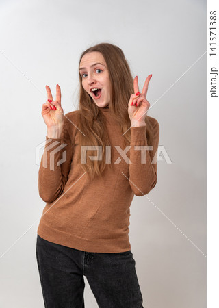 Young woman smiling with an excited expression, wearing a brown turtleneck and dark jeans, standing on a white background, making a V sign with both hands, representing success and happiness Young woman smiling with an excited expression, wearing a brown turtleneck and dark jeans, standing on a white background, making a V sign with both hands, representing success and happiness 141571388