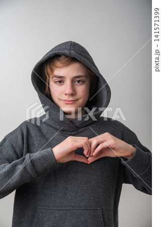 Young person in a grey hooded sweatshirt looks at camera and makes a heart with both hands, expressing love, kindness and gratitude in a casual studio portrait Young person in a grey hooded sweatshirt looks at camera and makes a heart with both hands, expressing love, kindness and gratitude in a casual studio portrait 141571399
