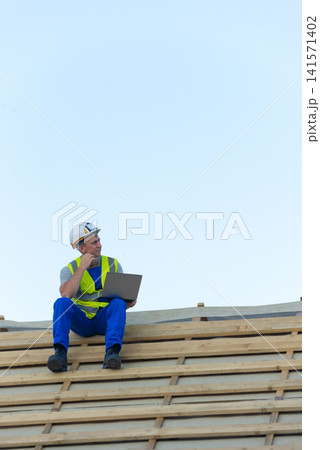 Construction worker in hard hat and safety vest sitting on new roof framework, holding a laptop and contemplating design plans against a clear blue sky Construction worker in hard hat and safety vest sitting on new roof framework, holding a laptop and contemplating design plans against a clear blue sky 141571402