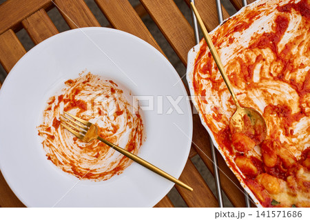 Empty plate with tomato sauce traces beside baking dish with leftover gnocchi Empty plate with tomato sauce traces beside baking dish with leftover gnocchi 141571686