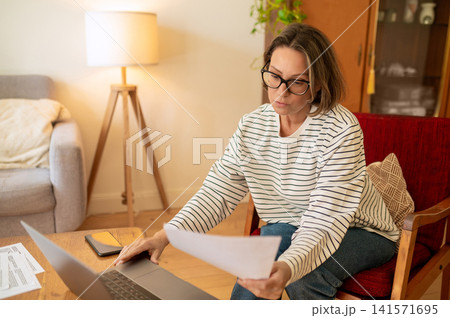 Middle aged female specialist analyzing paper near laptop 141571695