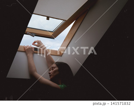Young woman reaching toward an open skylight window on a hot summer day. Lifestyle image representing heatwave, summer fatigue and fresh air. Young woman reaching toward an open skylight window on a hot summer day. Lifestyle image representing heatwave, summer fatigue and fresh air. 141571832