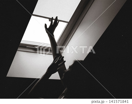 Silhouette of a woman reaching toward a bright skylight while shielding herself from intense light. Conceptual image representing photophobia, sensory sensitivity, migraine triggers and anxiety. Silhouette of a woman reaching toward a bright skylight while shielding herself from intense light. Conceptual image representing photophobia, sensory sensitivity, migraine triggers and anxiety. 141571841