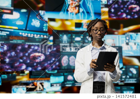 Researcher in lab coat analyzing human anatomy using tablet app in front of digital health dashboards backdrop. Female scientist uses device to project medical scans visualizations on videowall Researcher in lab coat analyzing human anatomy using tablet app in front of digital health dashboards backdrop. Female scientist uses device to project medical scans visualizations on videowall 141572481