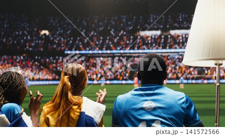 Football fans watch match on sofa with popcorn and beverages, interactive viewing experience making them feel close to stadium field. Immersive TV broadcast brings live sports venue atmosphere to home 141572566