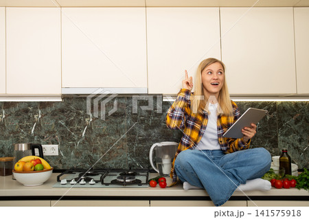 Cooking. Home cook sits on kitchen counter with tablet, joyfully exploring recipe Cooking. Home cook sits on kitchen counter with tablet, joyfully exploring recipe 141575918