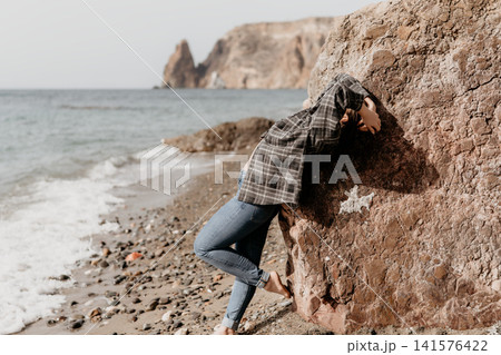 Woman beach rock leaning on a large boulder while barefoot on a pebble seashore with coastal cliffs in the background Woman beach rock leaning on a large boulder while barefoot on a pebble seashore with coastal cliffs in the background 141576422