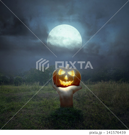 A hand rising from the ground holding a glowing carved pumpkin under a bright full moon in a spooky night field. Halloween and horror background A hand rising from the ground holding a glowing carved pumpkin under a bright full moon in a spooky night field. Halloween and horror background 141576439