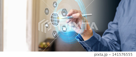 A man in a blue shirt uses a stylus to select icons on a glowing futuristic digital HUD interface displaying global connection, security, and smart home symbols. Concept of Remote working A man in a blue shirt uses a stylus to select icons on a glowing futuristic digital HUD interface displaying global connection, security, and smart home symbols. Concept of Remote working 141576555