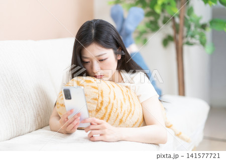 ソファでうつ伏せになって不安な表情でスマートフォンを見る若い女性 ソファでうつ伏せになって不安な表情でスマートフォンを見る若い女性 141577721