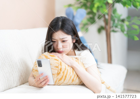 ソファでうつ伏せになって不安な表情でスマートフォンを見る若い女性 141577723