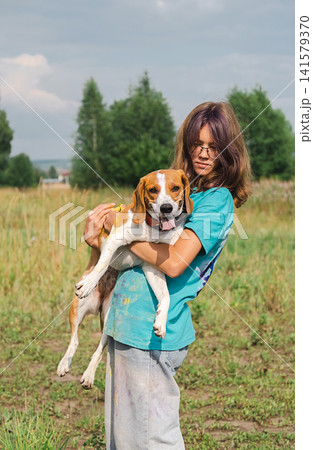 Young teenager girl holding cute beagle dog in meadow field. Happy child with pet enjoying nature outdoor. Young teenager girl holding cute beagle dog in meadow field. Happy child with pet enjoying nature outdoor. 141579370