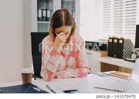 Young professional woman suffering from headache while analyzing financial charts and graphs on computer, experiencing work related stress in modern office Young professional woman suffering from headache while analyzing financial charts and graphs on computer, experiencing work related stress in modern office 141579470