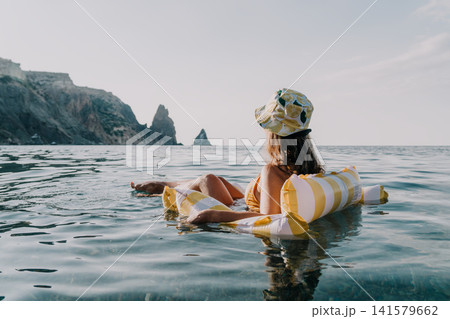 Woman Beach Inflatable Relaxing on a striped inflatable float in the ocean near a rocky coastline, wearing a hat and enjoying the sun. Woman Beach Inflatable Relaxing on a striped inflatable float in the ocean near a rocky coastline, wearing a hat and enjoying the sun. 141579662