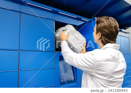 Man retrieving a package from a blue parcel locker. Fast ecommerce contactless delivery, pickup, and logistics technology at a self-service terminal. Man retrieving a package from a blue parcel locker. Fast ecommerce contactless delivery, pickup, and logistics technology at a self-service terminal. 141580443