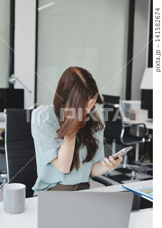 Asian businesswoman having a stressful phone call, holding a pen, and looking troubled while working on her laptop with financial documents and a calculator on her office desk Asian businesswoman having a stressful phone call, holding a pen, and looking troubled while working on her laptop with financial documents and a calculator on her office desk 141582674