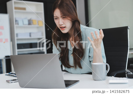Asian businesswoman having a stressful phone call, holding a pen, and looking troubled while working on her laptop with financial documents and a calculator on her office desk Asian businesswoman having a stressful phone call, holding a pen, and looking troubled while working on her laptop with financial documents and a calculator on her office desk 141582675